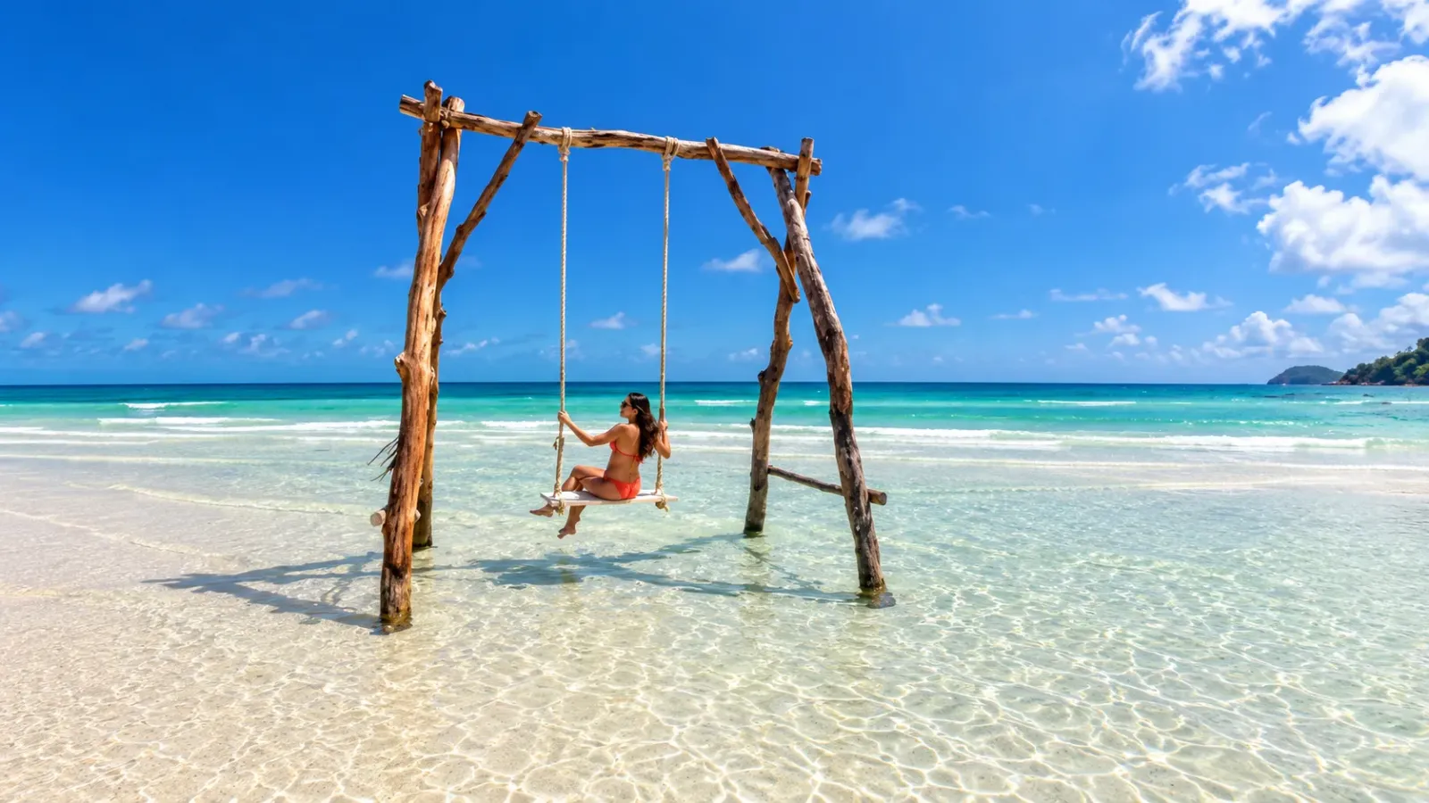 Du khách ngồi trên xích đu gỗ cạnh bãi biển, phía sau là mặt nước xanh và cầu gỗ