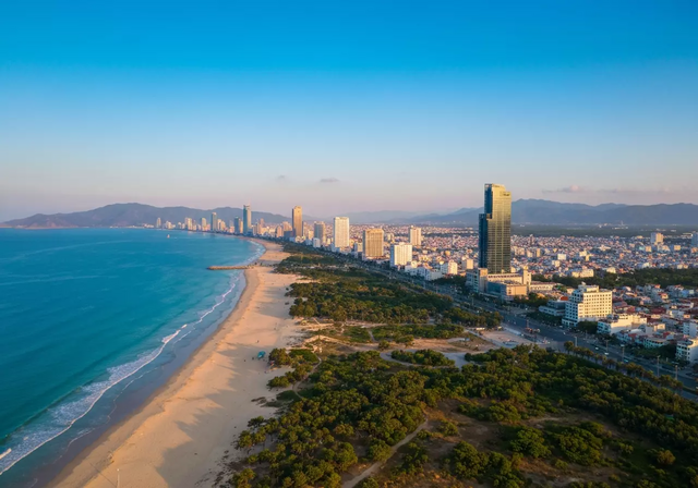 Toàn cảnh thành phố Nha Trang và bờ biển từ trên cao vào buổi hoàng hôn, với bầu trời trong xanh, biển ngọc bích êm đềm, bãi cát dài và đường chân trời hiện đại, tạo nên một khung cảnh yên bình