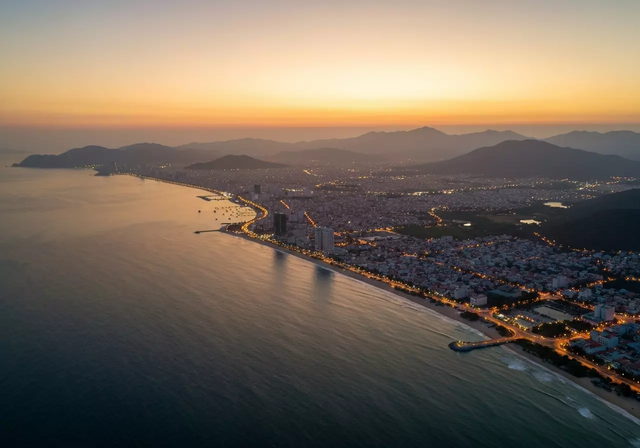 Toàn cảnh thành phố Nha Trang lúc hoàng hôn nhìn từ trên cao, đường bờ biển uốn lượn, những ánh đèn thành phố bắt đầu lung linh, núi non hùng vĩ phía sau và đại dương tĩnh lặng, tất cả chìm trong ánh sáng vàng ấm áp của buổi chiều tà, tạo nên một khung cảnh yên bình.