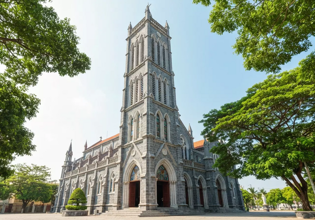 Nhà Thờ Đá Nha Trang với kiến trúc Gothic uy nghi, mặt tiền đá xám, tháp chuông cao vút và những ô cửa kính màu sặc sỡ, bao quanh bởi cây xanh, tạo nên một khung cảnh trang nghiêm dưới ánh sáng ban ngày